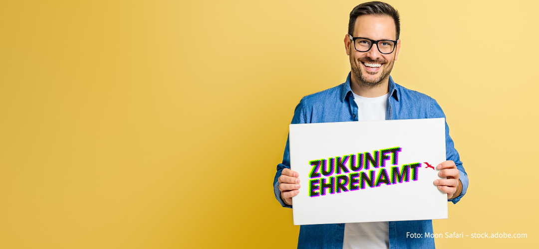 Ein Mann vor gelbem Hintergrund hält ein Plakat mit dem Logo 
