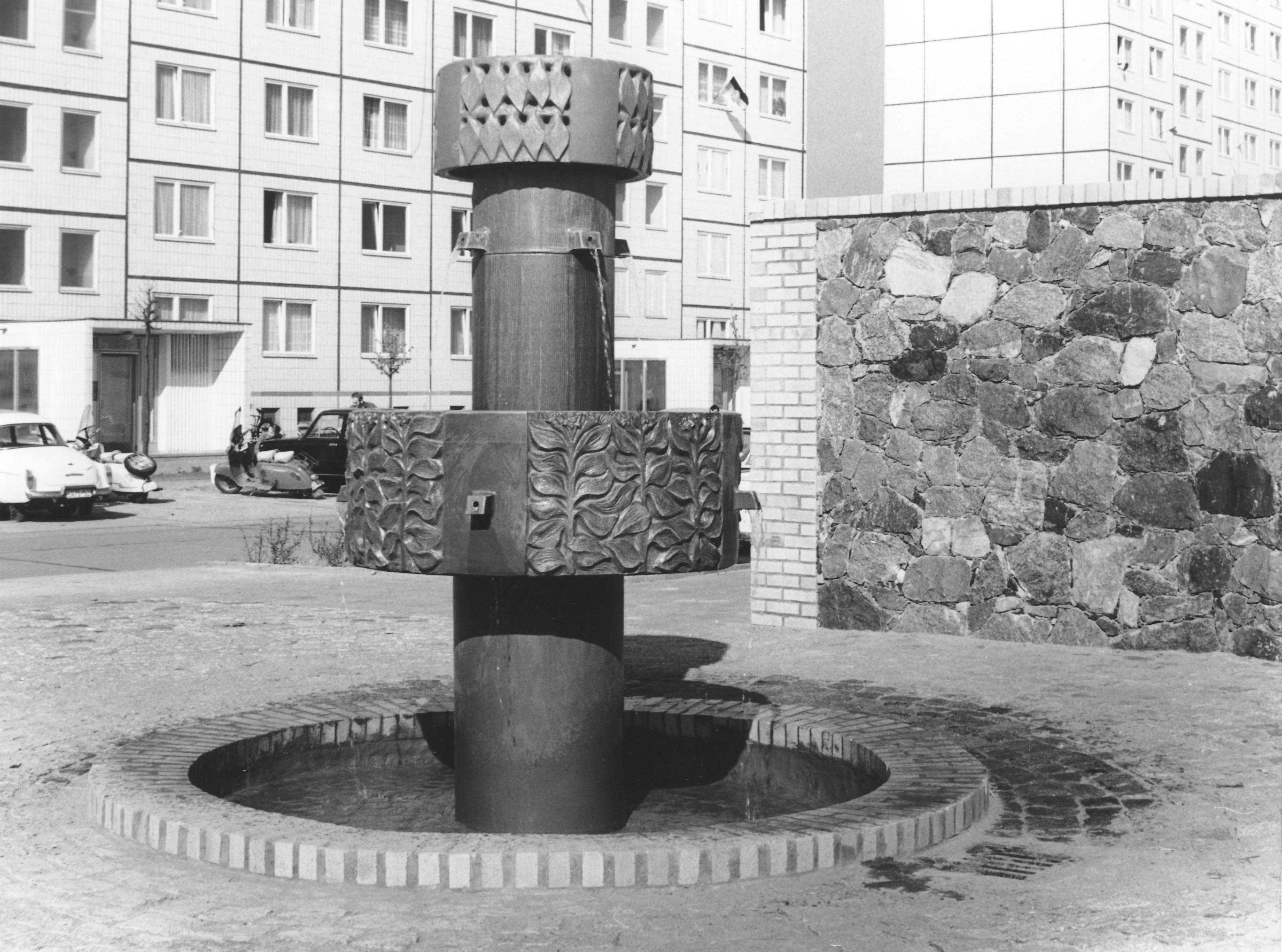 Schwarz-weiss Foto. Im Vordergrund steht ein Brunnen. Der ist aus Keramik und sieht wie eine Säule mit einem breiten Ring in der Mitte. Im Hintergrund sind Plattenbauten.