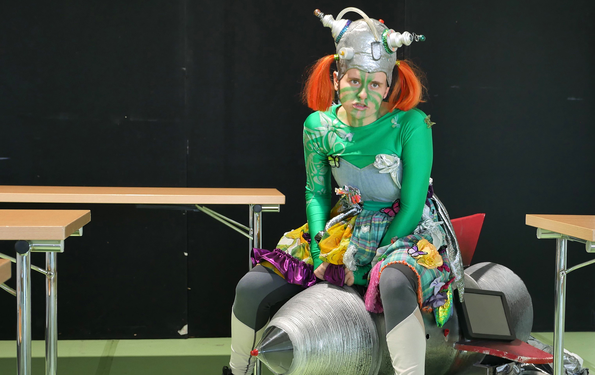 Foto: dSchauspielerin als Außerirdische verkleidet sitzt auf einer Rakete im Klassenzimmer