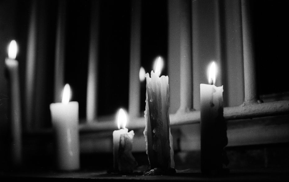 viele brennende Kerzen in der Dunkelheit vor einem vergitterten Fenster 