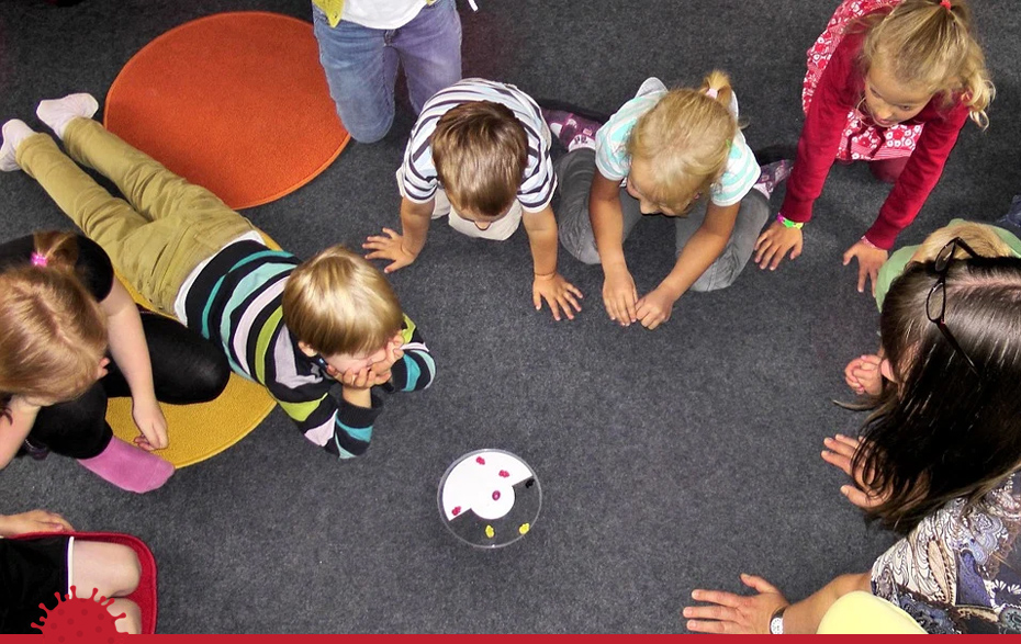 Foto: Kinder liegen bzw. knieen im Kreis auf dem Boden und verfolgen ein Spiel.