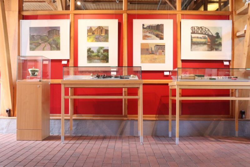 Mehrere Bilder von Scheunen im Foyer des Tabakmuseums. Die Bilder hängen auf einer roten Wand. zu sehen ist noch eine Glasvitrine der Dauerausstellung mit Pfeifen.