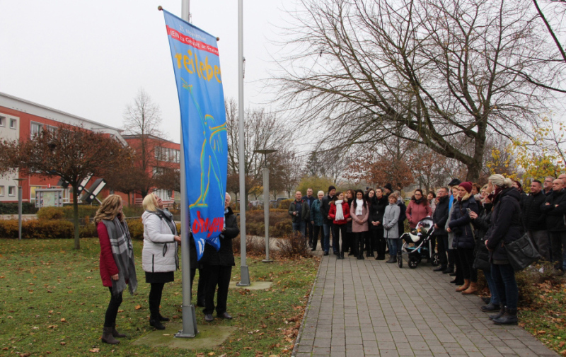 Foto: Die Flagge wird hochgezogen im Beisein zahlreicher Frauen und Männer.