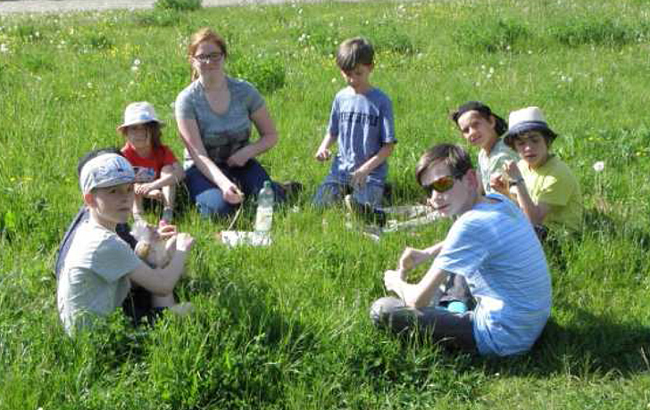 Foto: Kinder und eine Betreuerin sitzen im Kreis auf einer Wiese.